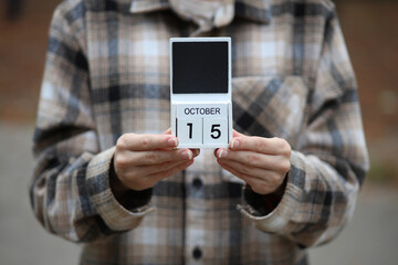 Woman in warm shirt holds calendar with date October 15 outdoors in autumn park