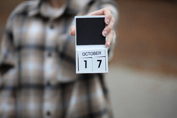 Woman in warm shirt holds calendar with date October 17 outdoors in autumn park