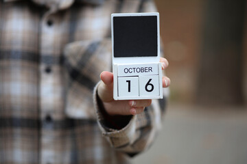 Woman in warm shirt holds calendar with date October 16 outdoors in autumn park