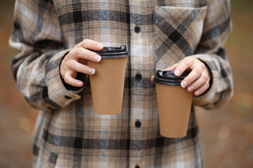 Woman in warm shirt holds cardboard cups for hot drink (coffee, cup) outdoors in autumn park