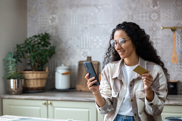 Young beautiful woman in kitchen with phone and bank credit debit card in hands, smiling Hispanic woman chooses gifts and products in online store, online shopping remotely from home. - obrazy, fototapety, plakaty