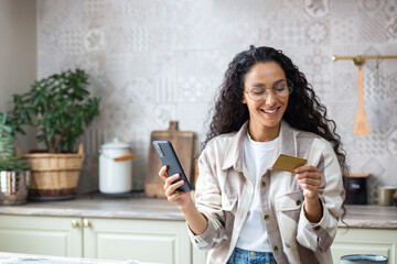 Young beautiful woman in kitchen with phone and bank credit debit card in hands, smiling Hispanic...