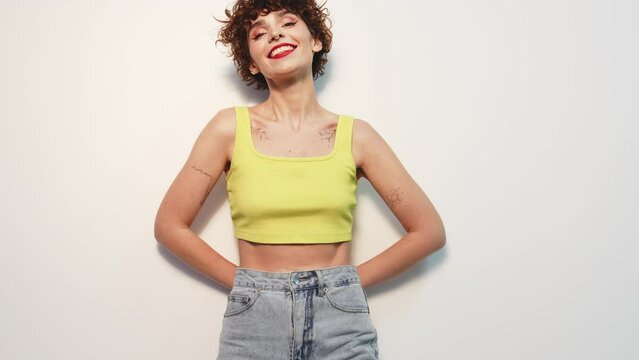 Young beautiful smiling female in trendy summer yellow tank top. Carefree woman posing near white wall in studio with curly hairstyle. Positive model having fun, going crazy