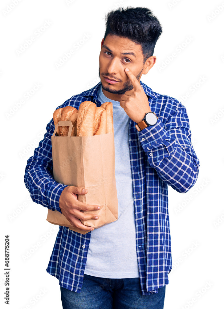 Wall mural Handsome latin american young man holding paper bag with bread pointing to the eye watching you gesture, suspicious expression