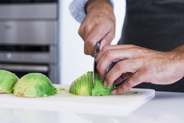Closeup of Crop unrecognizable chef in apron slicing avocado with knife on cutting board