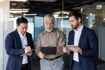 Three young businessmen of different nationalities are standing in the office, holding a tablet and...