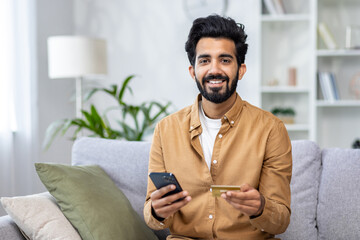 Portrait of a young Indian man sitting on the sofa at home, holding a credit card and a mobile phone, smiling and looking at the camera. - obrazy, fototapety, plakaty