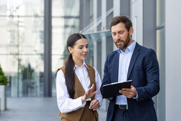 A young business man and a business woman stand outside the office center and discuss the documents...