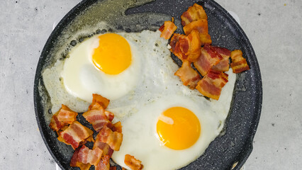 Two fried eggs and crispy fried bacon slices close-up on a hot frying pan.