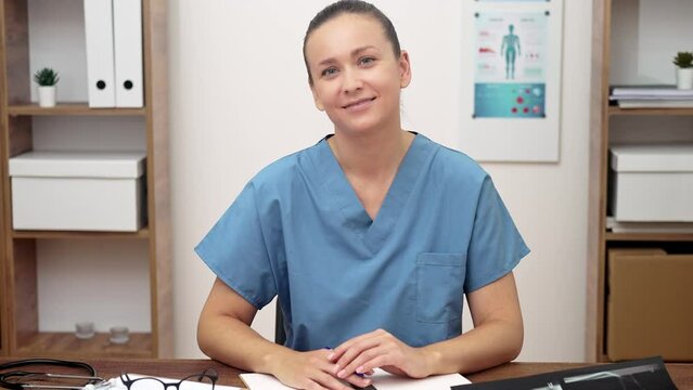 doctor in a blue uniform looking to camera smiling, medical documents and test results on her desk.