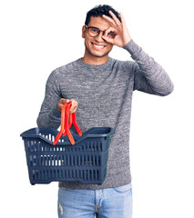 Hispanic handsome young man holding supermarket shopping basket smiling happy doing ok sign with...