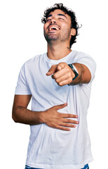 Hispanic young man with beard wearing casual white t shirt laughing at you, pointing finger to the camera with hand over body, shame expression