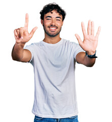 Hispanic young man with beard wearing casual white t shirt showing and pointing up with fingers number seven while smiling confident and happy.