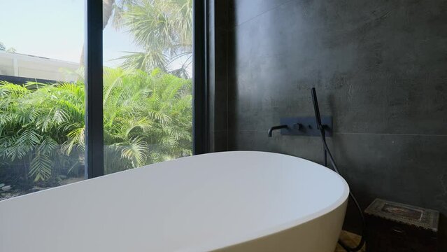 White modern bathtub next to panoramic window with wonderful view on tropical garden. Upper-class fashionable bathroom covered with black ceramic tiles on walls