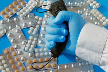 A hand in a blue medical glove crushed a black medical mask against the background of scattered...