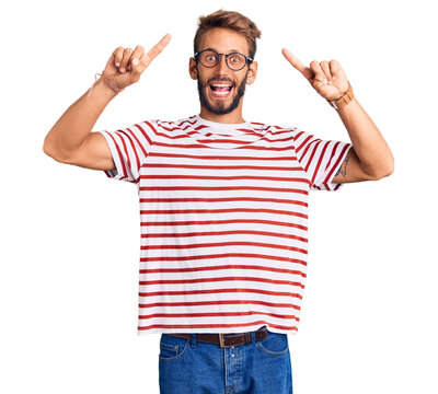 Handsome blond man with beard wearing casual clothes and glasses smiling amazed and surprised and pointing up with fingers and raised arms.