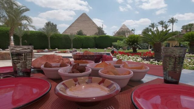 Egyptian food is on a table. Restaurant. Arabic cuisine. Pyramids of Giza is on the background. Luxury hotel terrace with palms and view on a Great Pyramids. Woman taking a photo.