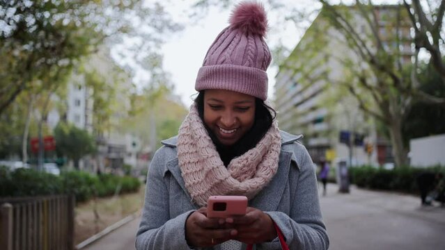 Front view of smiling young African American woman using smartphone while walking in city street. Female having fun looking his mobile through a garden area on a winter christmas day. 