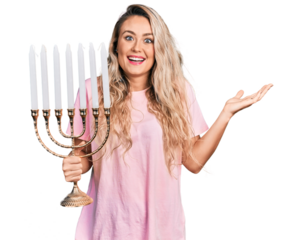 Young blonde woman holding menorah hanukkah jewish candle celebrating achievement with happy smile and winner expression with raised hand