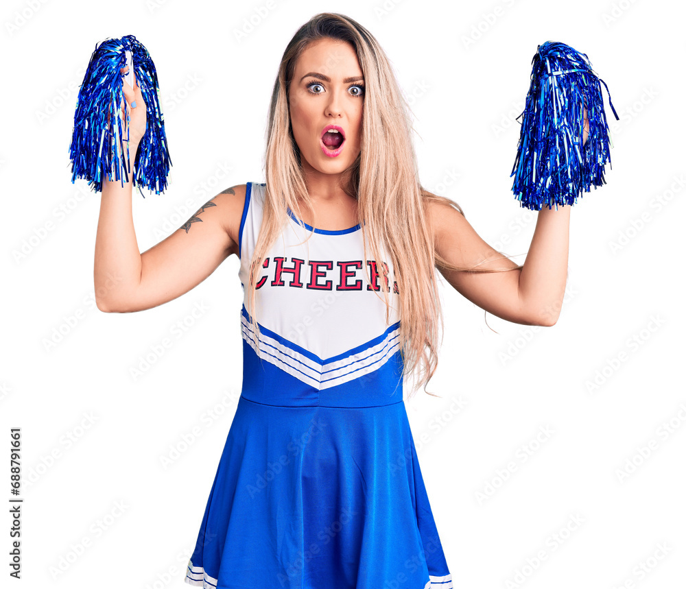 Poster young beautiful blonde woman wearing cheerleader uniform holding pompom scared and amazed with open 