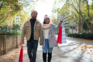 Beautiful young couple interracial in love enjoying winter holiday season in the city streets, taking a walk, pointing somewhere, hugging and holding gift bags while celebrating Christmas outdoors