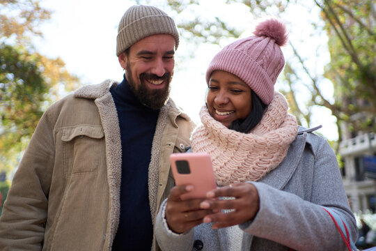 Beautiful young couple interracial in love using mobile phone enjoying winter holiday season in the city streets, walking and hugging and holding gift bags while celebrating Christmas outdoors