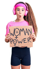 Cute hispanic child girl wearing gym clothes and using headphones holding woman power banner scared and amazed with open mouth for surprise, disbelief face