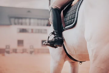 Fotobehang A rider in leather boots sits on a white horse in the saddle. Sports equipment and stirrups. Equestrian sports and horse riding. ©  Valeri Vatel