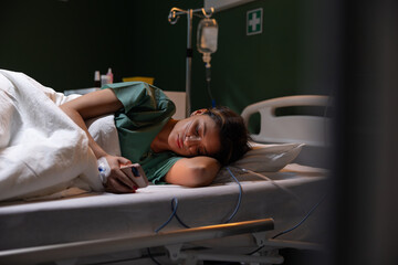 Young girl lies in the hospital room with a drip in her hand and a catheter, passing the time by playing games on her phone.
