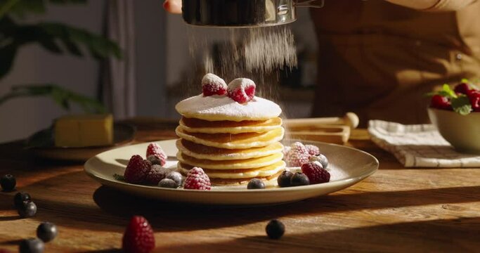 Sprinkling sugar powder on tasty pancake, slow motion. Classic flavors of American breakfast with pancakes beautifully set against the backdrop of cozy kitchen. Advertising. Pleasant family traditions