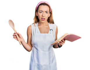 Young caucasian woman wearing professional baker apron reading cooking recipe book in shock face, looking skeptical and sarcastic, surprised with open mouth
