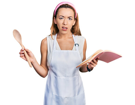 Young caucasian woman wearing professional baker apron reading cooking recipe book in shock face, looking skeptical and sarcastic, surprised with open mouth