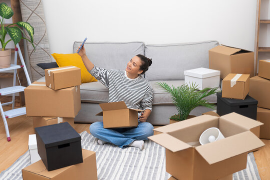 Woman takes selfies with box close to sofa in modern house waiting for lovely boyfriend from work to help sort out things needed to decorate contemporary cottage house