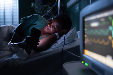African patient in hospital bed lies on her side, engrossed in a video call on the phone.