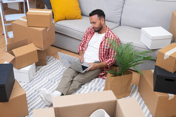 Young brunette guy sits with laptop among boxes in new flat checking availability of decorations...