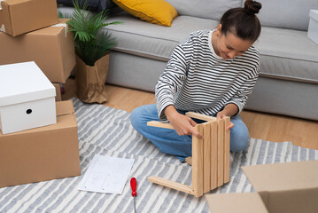 Cheerful woman sits with wooden furniture with positive expression on fabric carpet near sofa among cardboard boxes in spacious premise after receiving keys from realtor