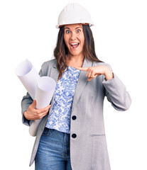 Young beautiful brunette woman wearing architect hardhat holding blueprint smiling happy pointing with hand and finger