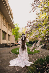 Brunette girl with long hair with big bow in long dress with train standing in garden of blooming...