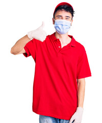 Young hispanic man wearing delivery uniform and medical mask smiling happy and positive, thumb up doing excellent and approval sign