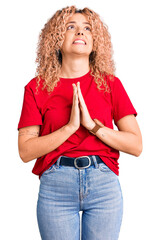 Young blonde woman with curly hair wearing casual red tshirt begging and praying with hands together with hope expression on face very emotional and worried. begging.