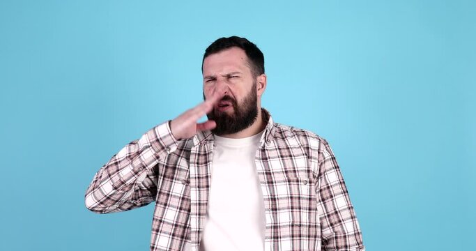 Caucasian bearded man isolated over blue background smelling something stinky and disgusting, intolerable smell, holding breath with fingers on nose. Bad smells concept.