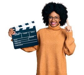 Young african american woman holding video film clapboard smiling with an idea or question pointing...