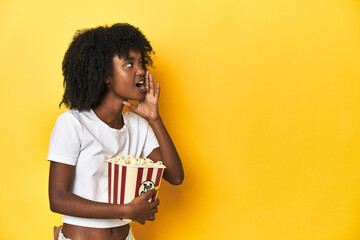 Teen girl with popcorn, cinema concept on yellow backdrop shouting and holding palm near opened mouth.