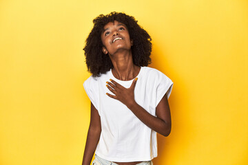 Teen girl in white tank top, yellow studio background laughs out loudly keeping hand on chest.