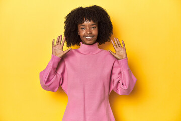 Teen girl in pink sweatshirt, yellow studio backdrop showing number ten with hands.