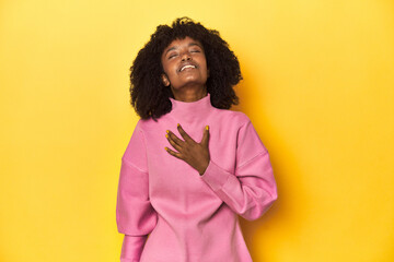 Teen girl in pink sweatshirt, yellow studio backdrop laughs out loudly keeping hand on chest.