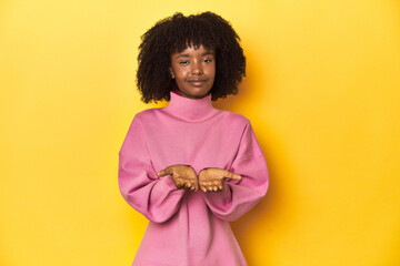 Obraz na płótnie Canvas Teen girl in pink sweatshirt, yellow studio backdrop holding something with palms, offering to camera.