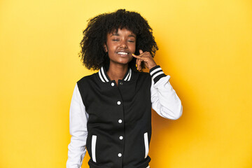 Teen girl in baseball jacket, yellow studio background showing a mobile phone call gesture with fingers.