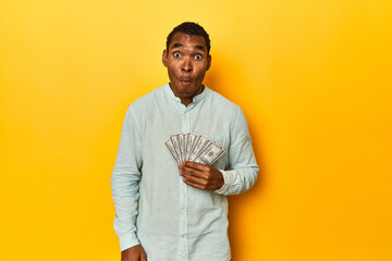 African American man with dollar bills, yellow studio, shrugs shoulders and open eyes confused. - obrazy, fototapety, plakaty