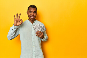 African American man with dollar bills, yellow studio, cheerful and confident showing ok gesture.